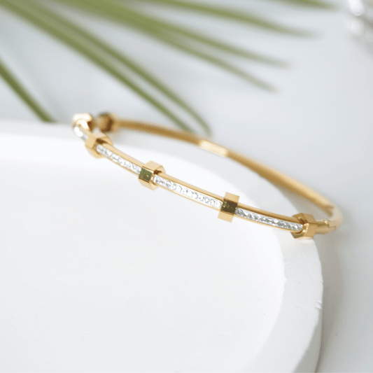 Gold bracelet with embedded stones on a white surface with green leaves in the background