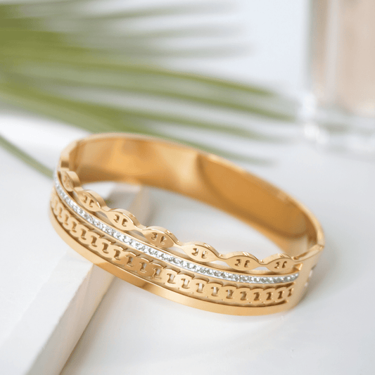 Gold bracelet with intricate design on a white surface with green leaves in the background