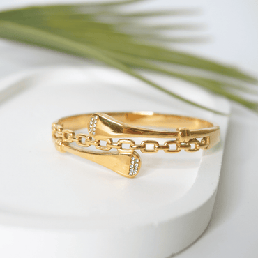 Gold bracelet with chain design on a white surface with green leaves in the background
