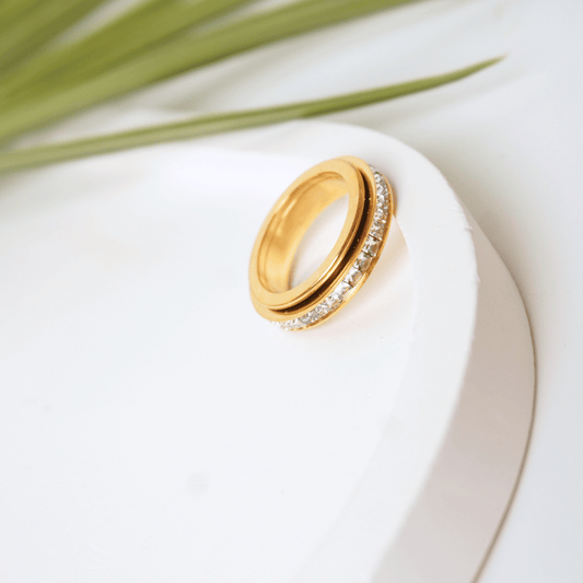 Gold ring with diamond accents on a white surface with green leaves in the background